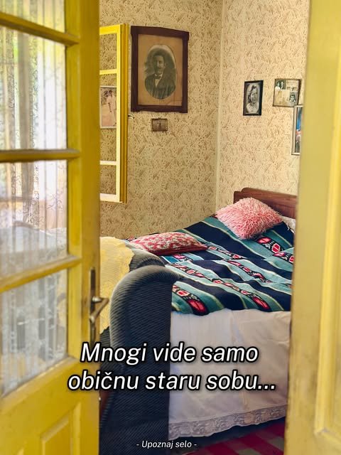 Stara seoska soba sa žutim prozorom, tkanim prekrivačem i uramljenim porodičnim fotografijama na zidu